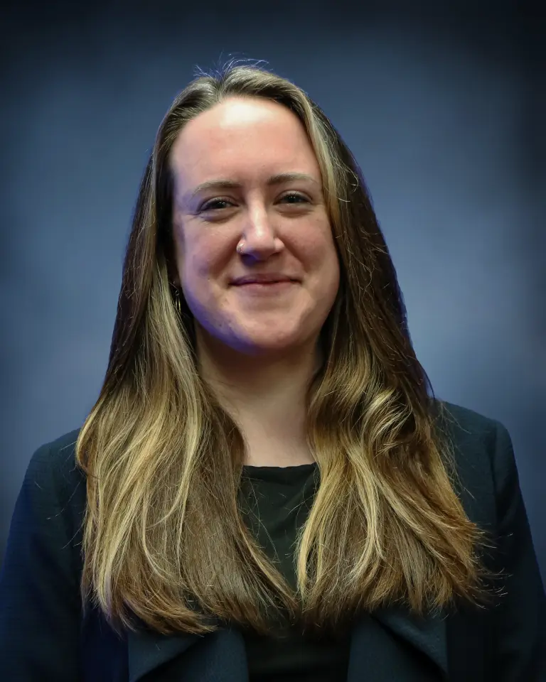 Ally McCabe, smiling, in a dark blazer against a plain grey backdrop.