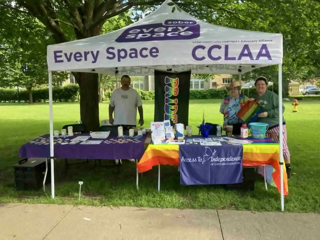 Three people stand behind the ATI and CANDLE table with pride flags decorating the booth.