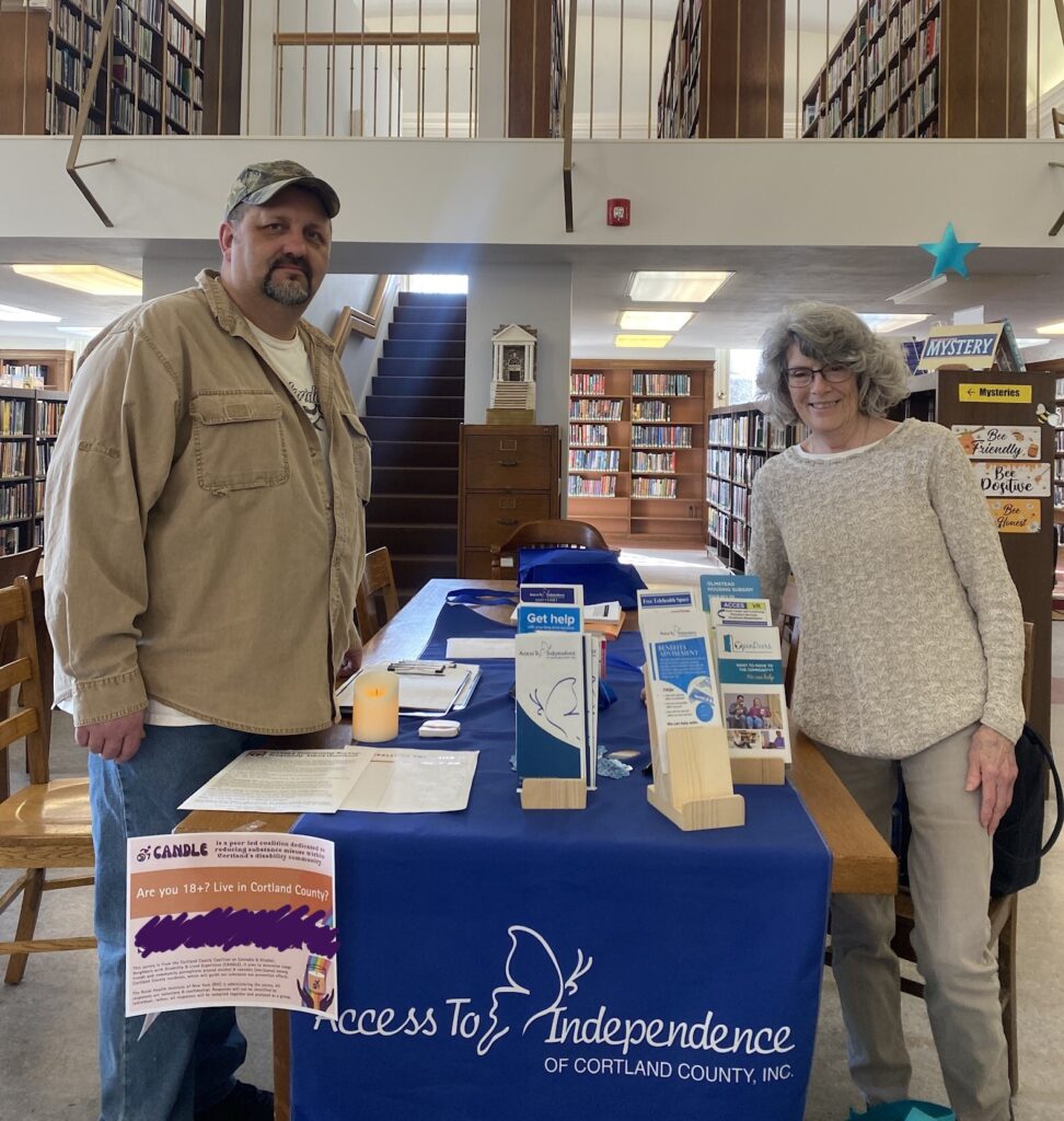 A white man and white woman smile at the camera as they stand by a CANDLE survey table at a library.