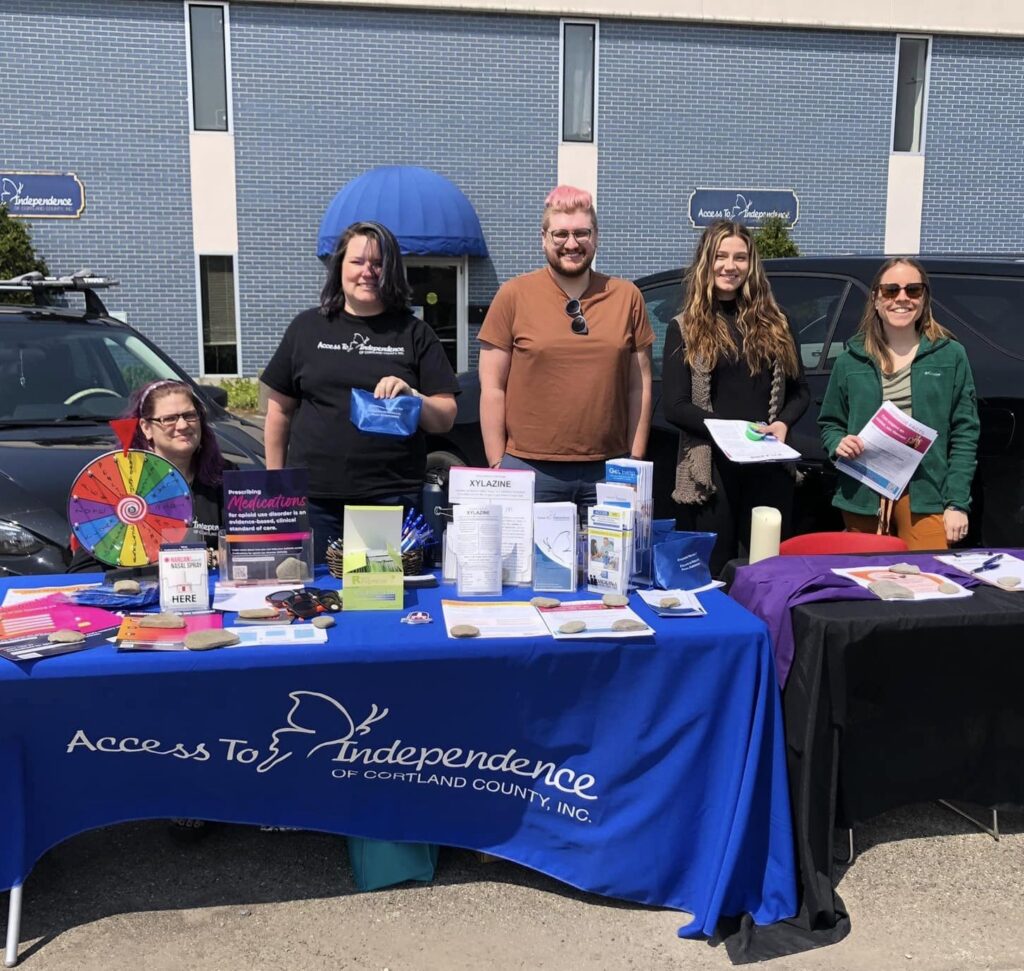 CANDLE staff and volunteers sit and stand behind an outreach table in ATI's parking lot. 